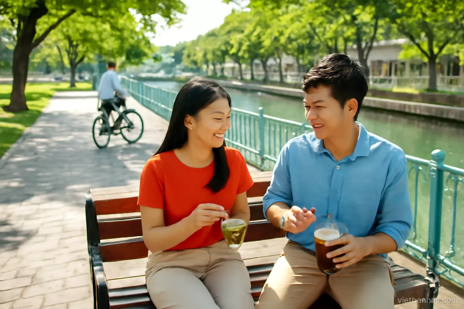 High-angle morning view of a shady canal-side path in Saigon, a young Vietnamese couple on a bench sharing sticky rice and iced coffee, a cyclist passing, fresh green trees.