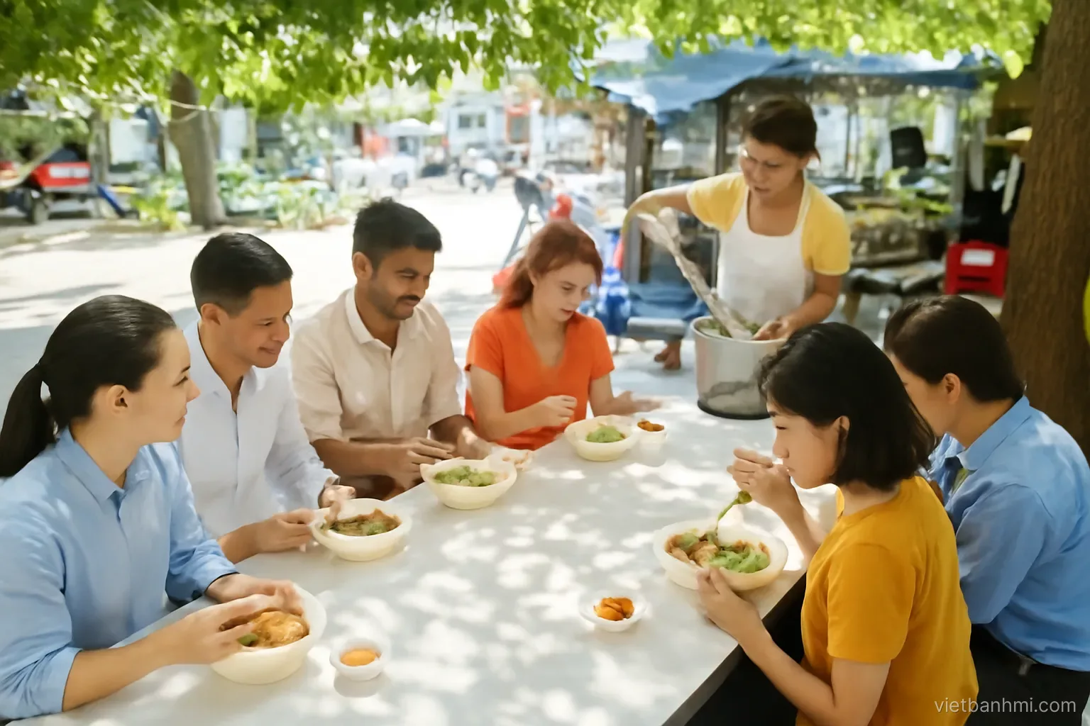 Vietnamese office workers and two mixed-ethnicity travellers sharing a long table under a shady tree at a busy lunchtime rice stall, cook ladling soup, high-angle wide shot, dappled midday light
